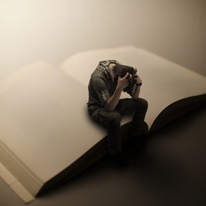 A man sitting on a giant book, both hands on his head and looking down on the floor, appearing to be in agony.