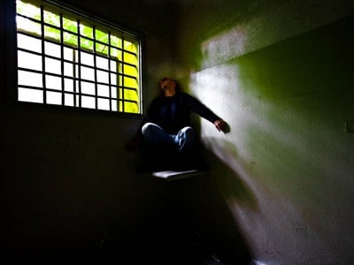 A man sitting on a high chair at a corner of the room, apearing to be in agony and looking at the window