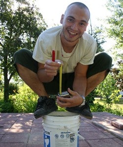 Carl squatting on a pail, taking a measuring tape and smiling.