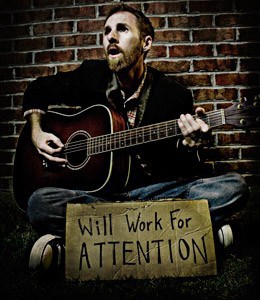 A man sitting on grass playiing a guitar, there's a card with the words: Will work for ATTENTION in front of him.