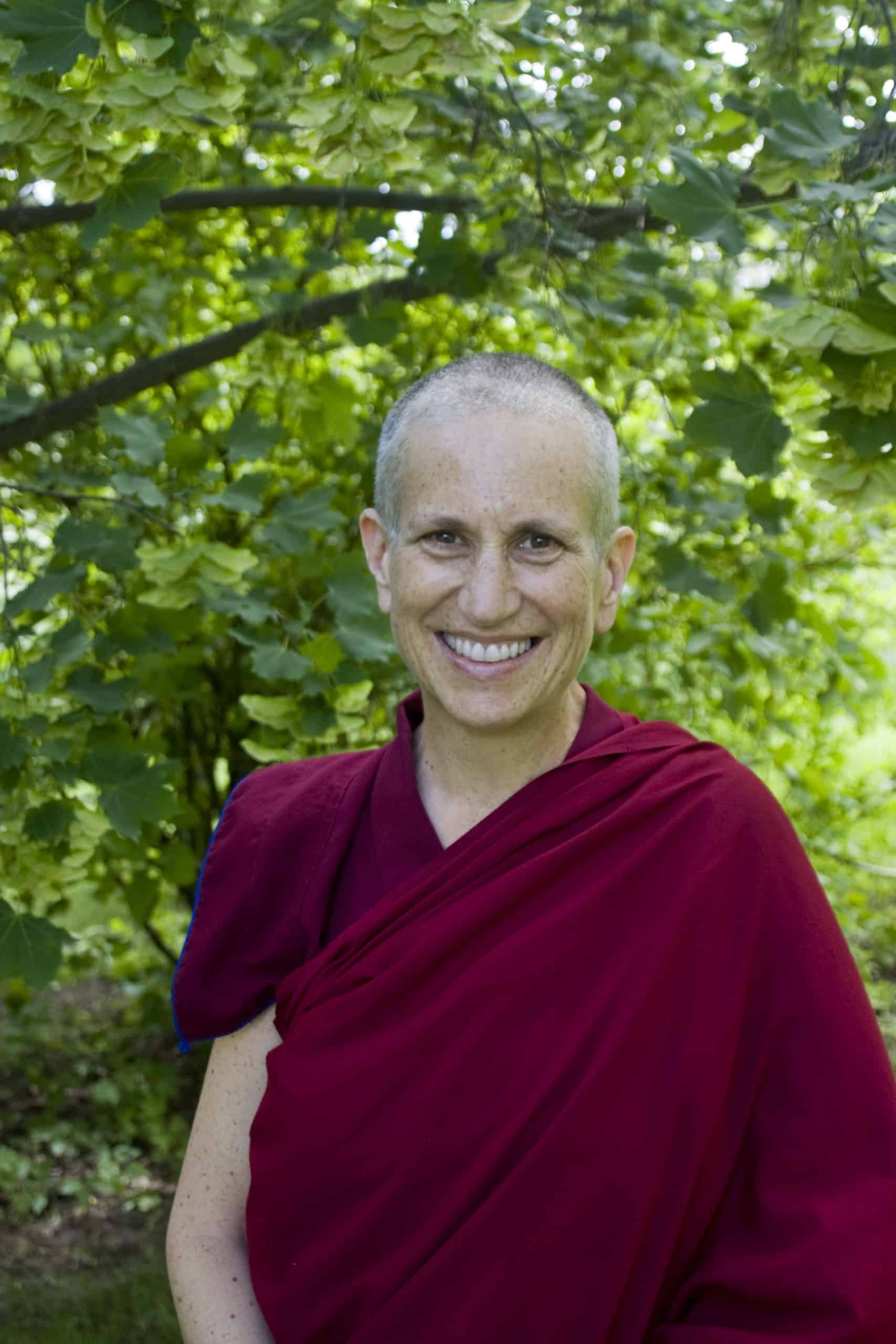 Venerable Chodron smiling, standing in front of a bright green tree.