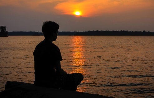 Silhouette of woman meditating by a lake, with sun setting in background.