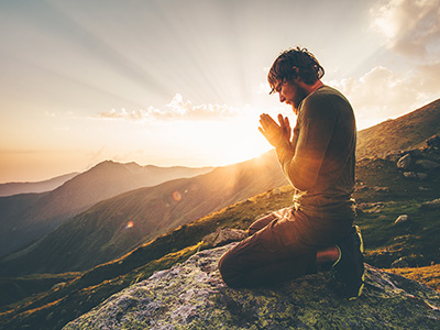 Man on a mountain at sunset, kneeling with palms together.