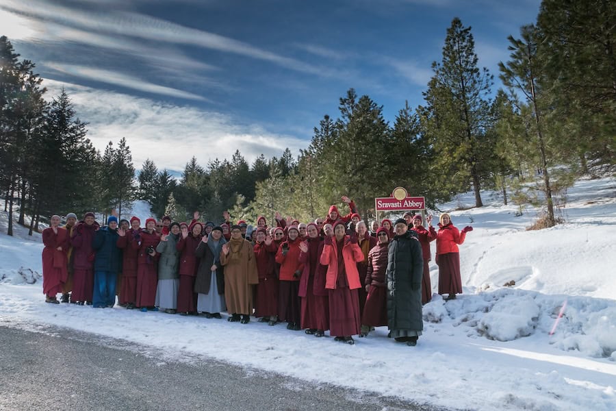 A group of nuns in winter clothing stand in front of the Sravasti Abbey sign.