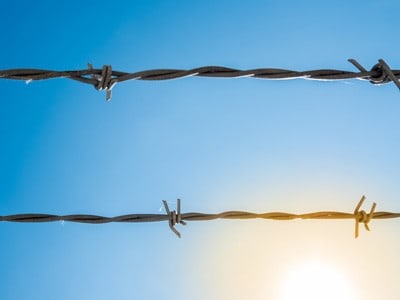 Two strands of barbed wire with blue sky in the background and sunlight.