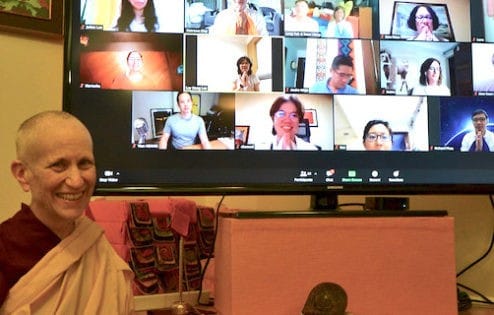 Venerable Chodron smiling while sitting in front of a large TV screen showing Zoom participants.