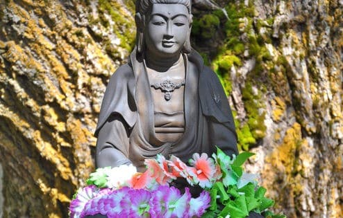 Kuan Yin statue on a pedestal with a flower garland in her lap under a tree.