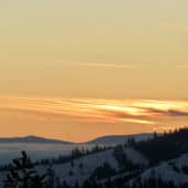 Orange sunset sky above mountains covered in trees and snow.