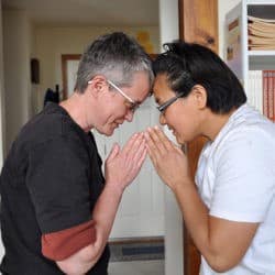 A Caucasian woman and an Asian woman touch foreheads and bow to each other with palms together.