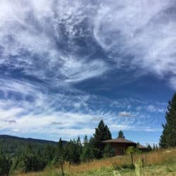 Clouds swirl in a blue sky over a green meadow and trees.