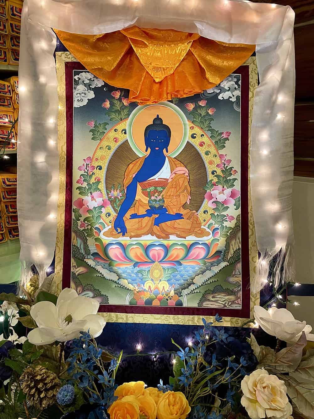 Medicine Buddha thangka surrounded by light and flower offerings.