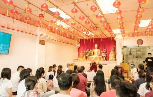 Venerable Chodron sitting in front of a large gold Buddha statue, teaching to a group of people.