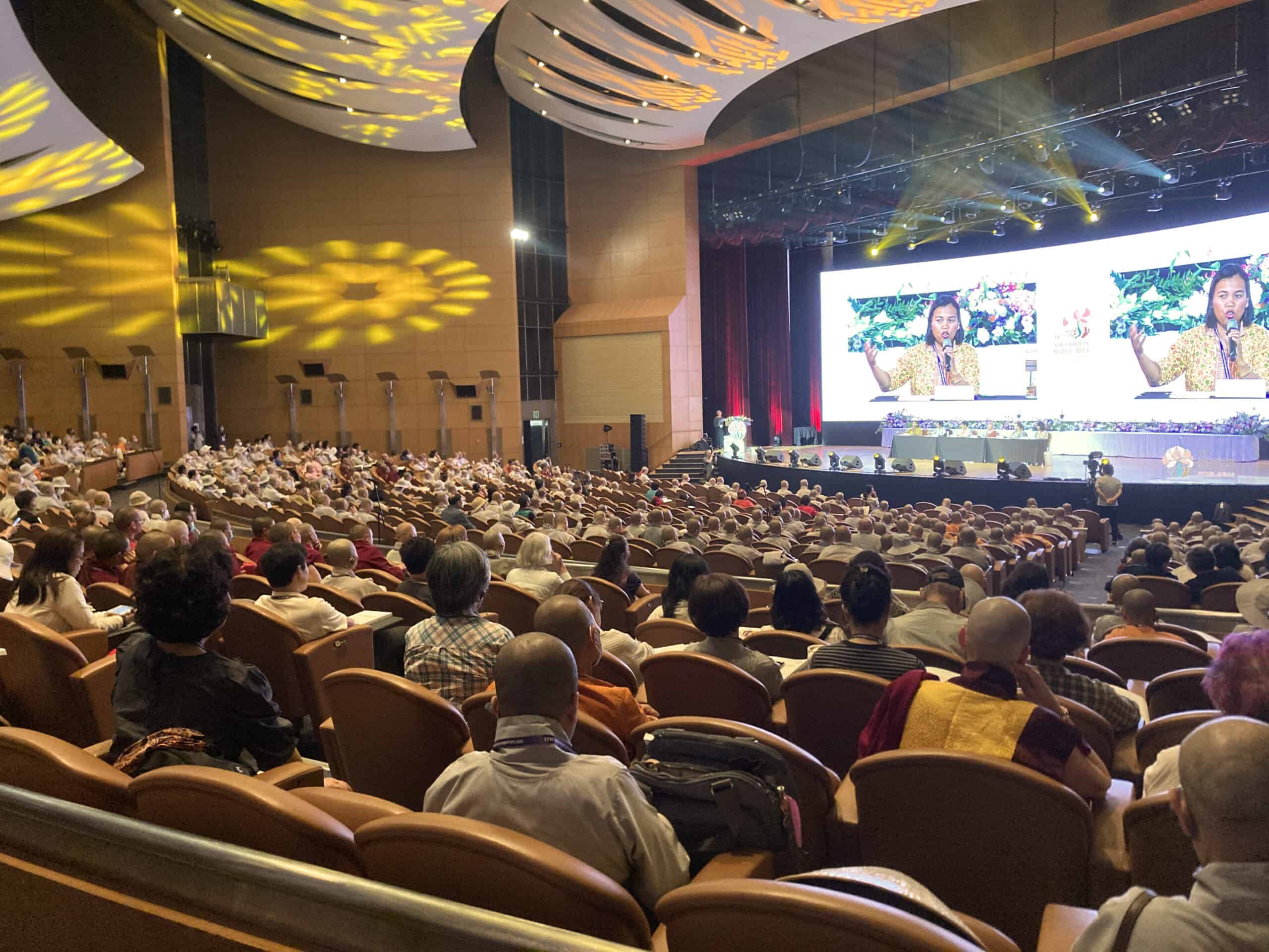 Group of people sitting in a crowded auditorium watching a presentation on a large screen.