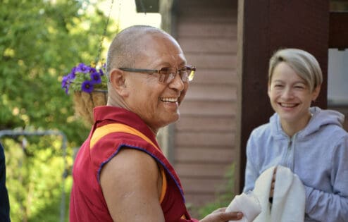 Geshe Tenzin Chodrak (Dadul Namgyal) smiling, a smiling student offering a khata in the background.