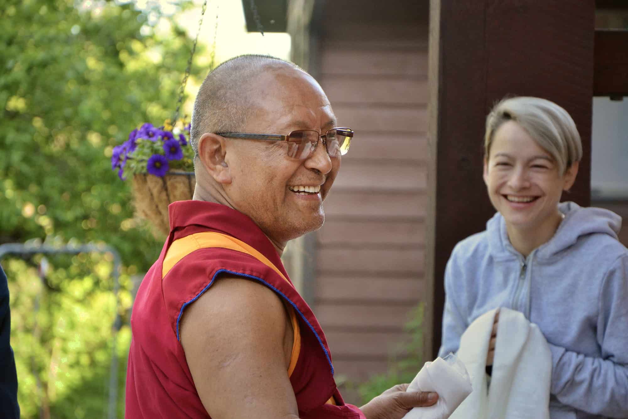 Geshe Tenzin Chodrak (Dadul Namgyal) smiling, a smiling student offering a khata in the background.